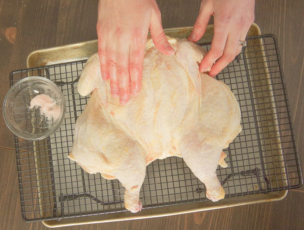 Hands rubbing salt on raw chicken sitting on a wire rack above roasting pan