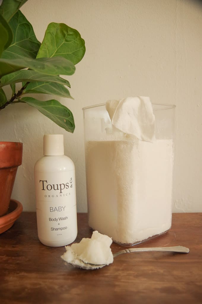 Homemade baby wipes in plastic container next to bottle of soap and spoonful of coconut oil