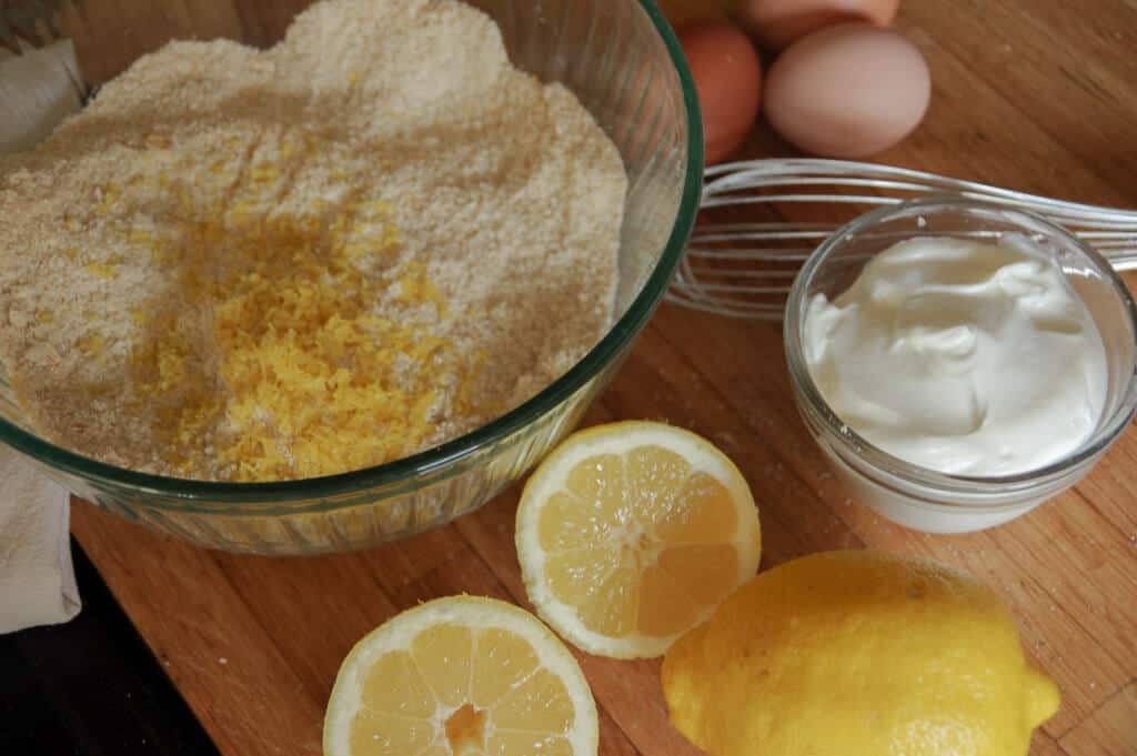 Bowl with flour and lemon zest in it. Lemons on counter and bowl of yogurt