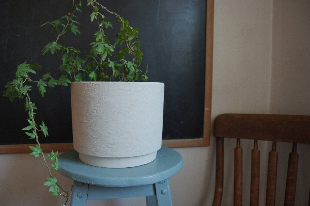 Clay planter with a vine inside on a blue stool next to a  chair and chalkboard