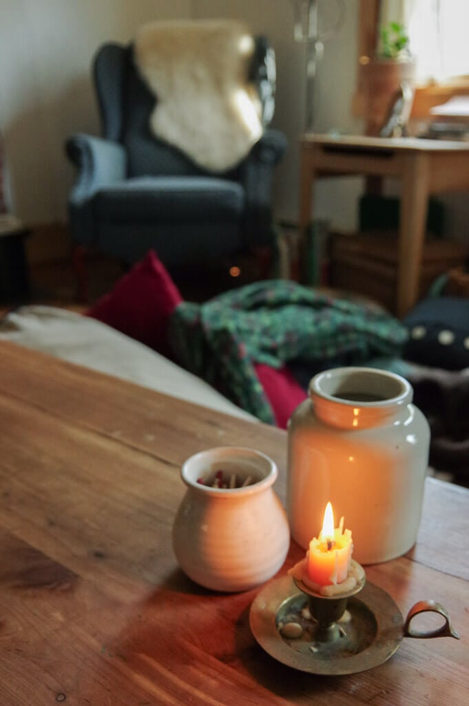 Lit Candle in front of two ceramic jars on wooden table with a blue chair in the background 